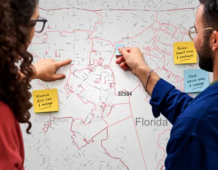 Two colleagues placing sticky notes on a redline carrier route wall map of ZIP code 32504 in Escambia County, Florida.