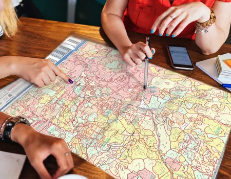 Hands pointing with a pen and a fingertip at a pastel carrier route map spread across a wooden table beside a phone.