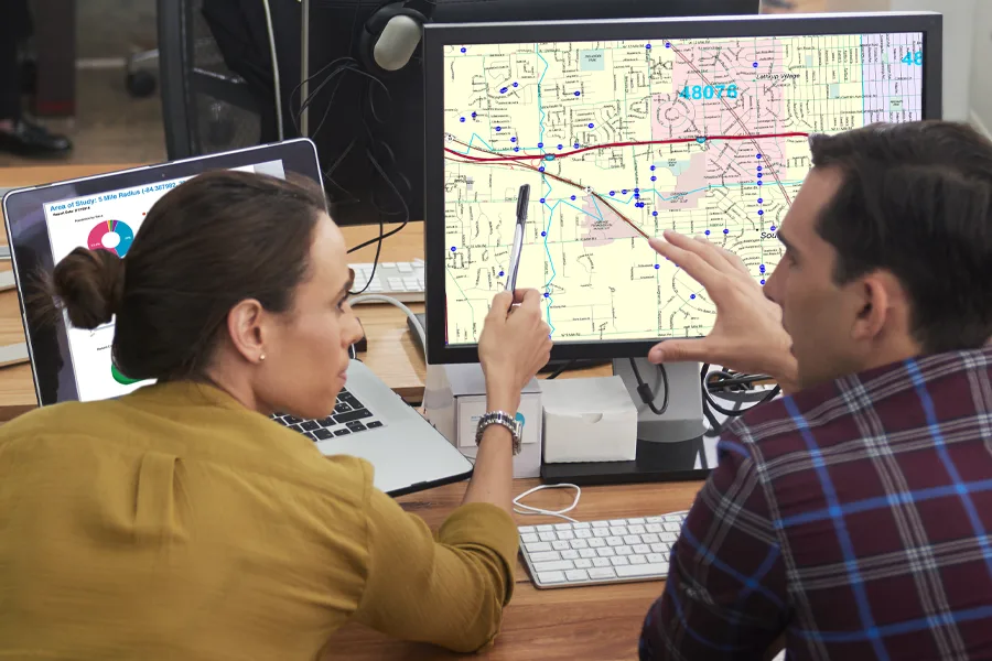 Two colleagues at a desk studying a street map on a monitor with delivery stops marked in blue, beside a laptop showing a 5-mile radius report.